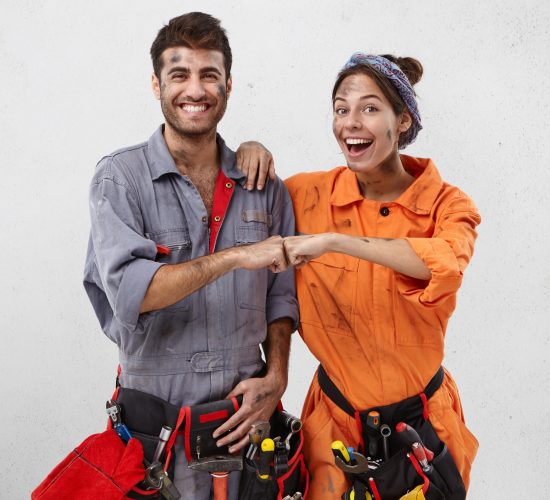 Happy team of service workers rejoice successful finishing work. Smiling male carpenter in special uniform with tool belt and overjoyed female colleague keep hands together, glad to have triumph